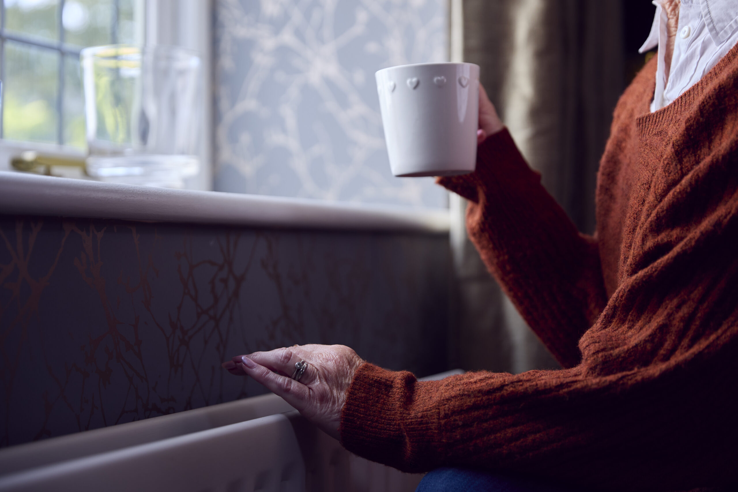 Senior Woman With Hot Drink Trying To Keep Warm By Radiator At Home In Cost Of Living Energy Crisis