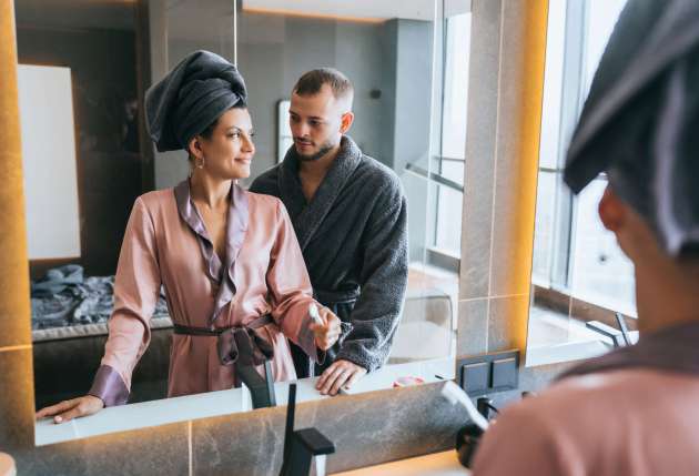 Couple in bathroom, woman smiling at mirror, man in bathrobe behind, cozy intimacy